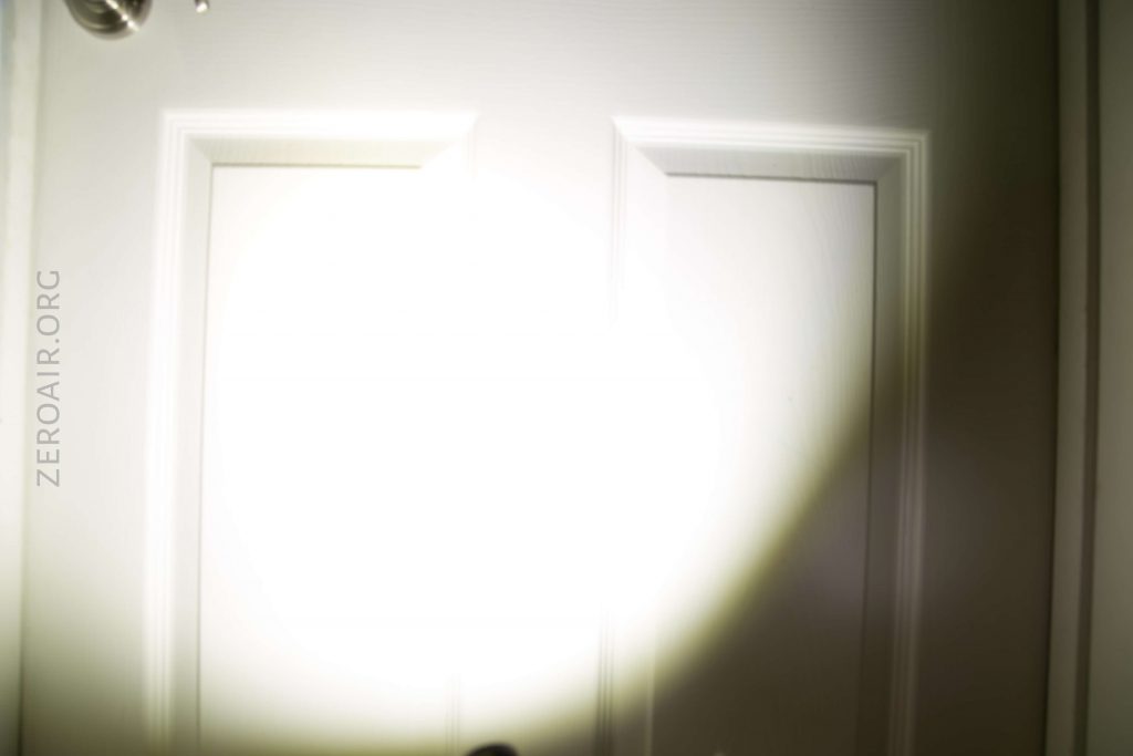 A white door with two raised rectangular panels is illuminated by a bright flashlight beam, creating a circular light spot at the center. The text “ZEROAIR.ORG” is visible along the left side of the image.