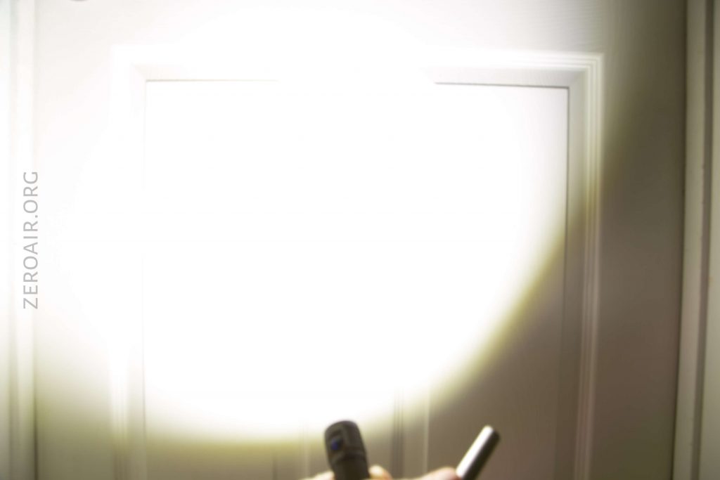 A bright flashlight beam illuminates a white door in a dark room. The hand holding the flashlight is partially visible at the bottom of the image. The website zeroair.org is faintly visible on the left side.