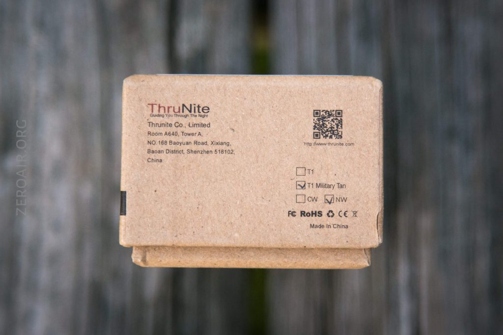 A brown cardboard box with a ThruNite logo and company information, including address, QR code, checkboxes for product color options, and certification symbols, is shown on a wooden surface.