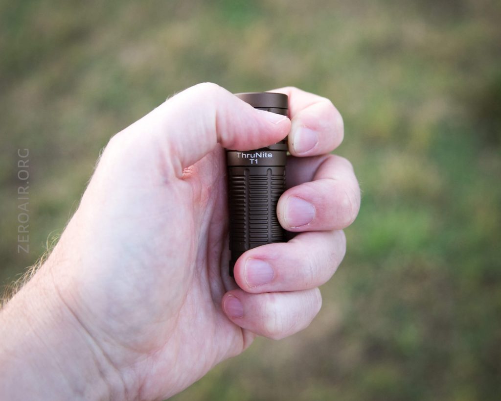 A hand holding a small, cylindrical black flashlight labeled ThruNite Ti against a blurred outdoor background. The fingers are wrapped around the flashlight, showing its compact size.