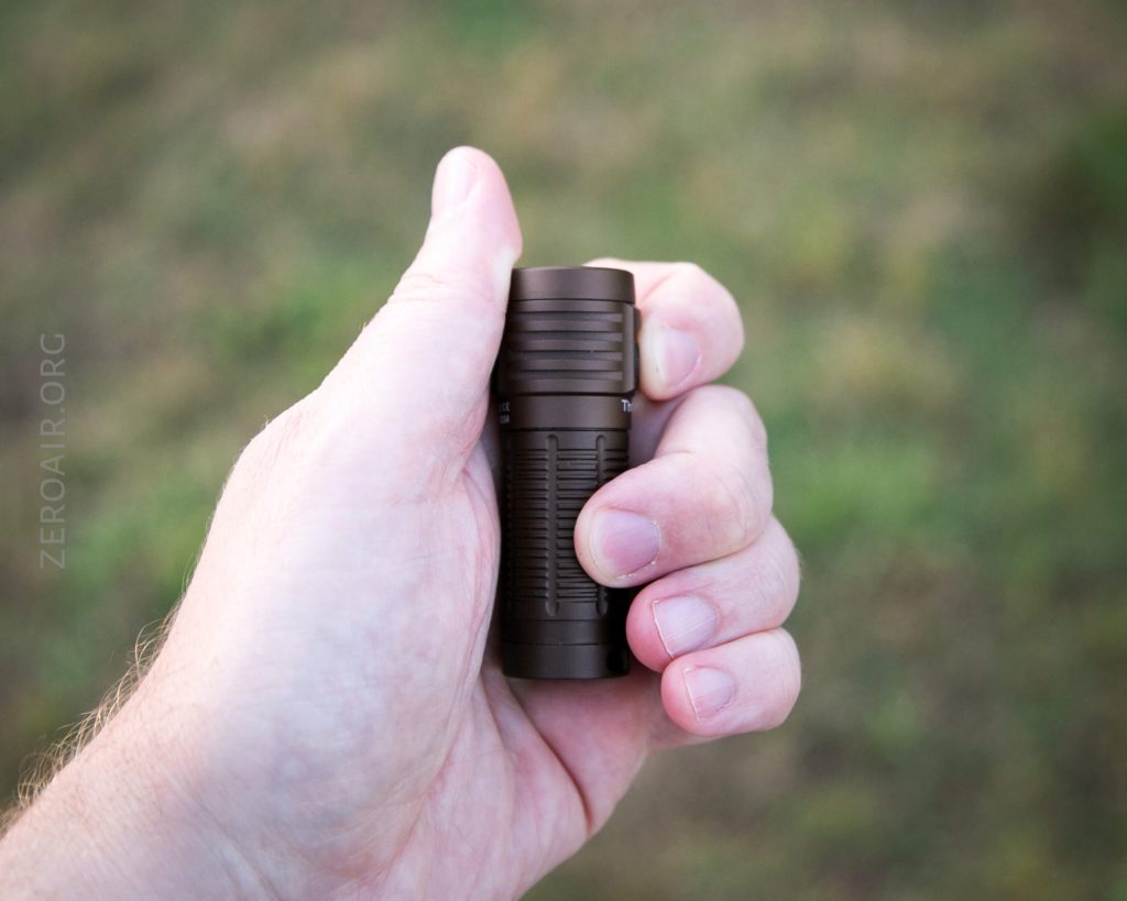 A light-skinned hand holding a small, dark-colored flashlight outdoors, with a blurred grassy background.