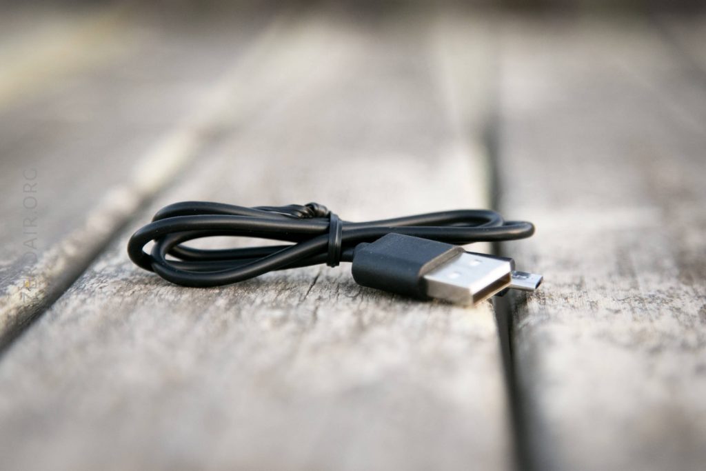 A black USB to micro USB charging cable lies coiled on a wooden surface, with the USB connector and micro USB connector both visible in the foreground.