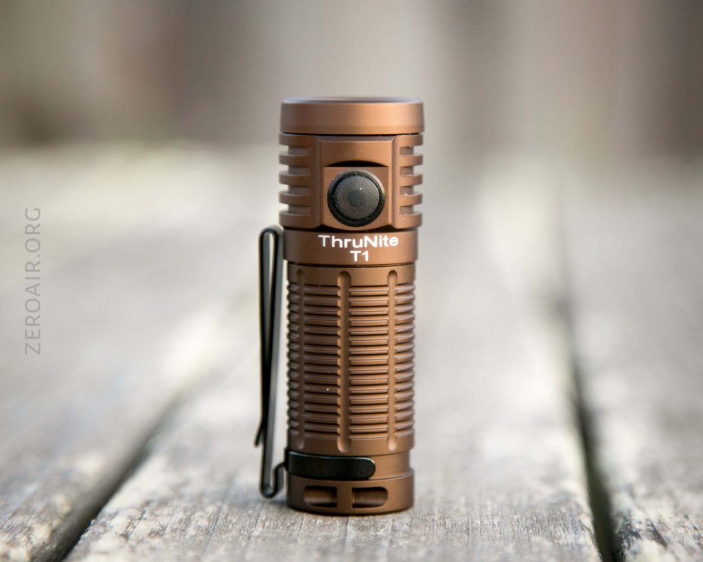 A bronze ThruNite T1 flashlight stands upright on a weathered wooden surface, with a side button and a pocket clip visible. The background is blurred.