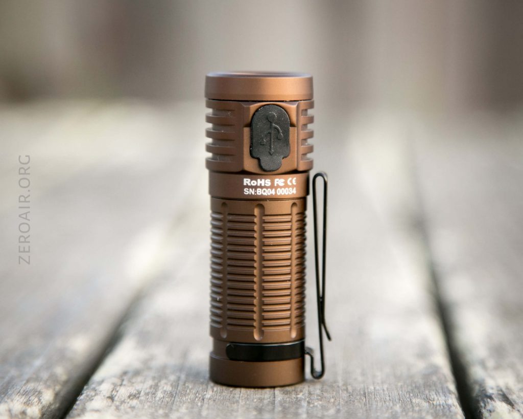 A small, cylindrical, brown flashlight with a ridged grip stands upright on a wooden surface. The flashlight has a side clip and visible compliance markings and serial number.