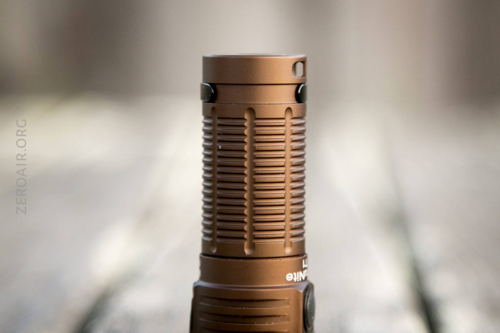 A close-up of a Thrunite T1 Military Tan Flashlight stands upright on a blurred, wooden surface. The brown, cylindrical flashlight features a textured grip and a matte finish.
