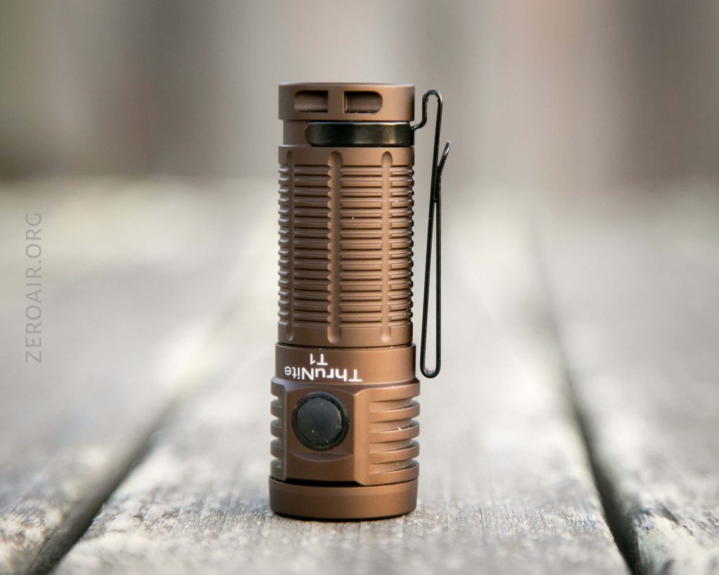 A compact Thrunite T1 Military Tan Flashlight stands upright on a wooden surface. The bronze-colored light features ribbed sides, a black side button, and a black pocket clip. The softly blurred background adds depth.