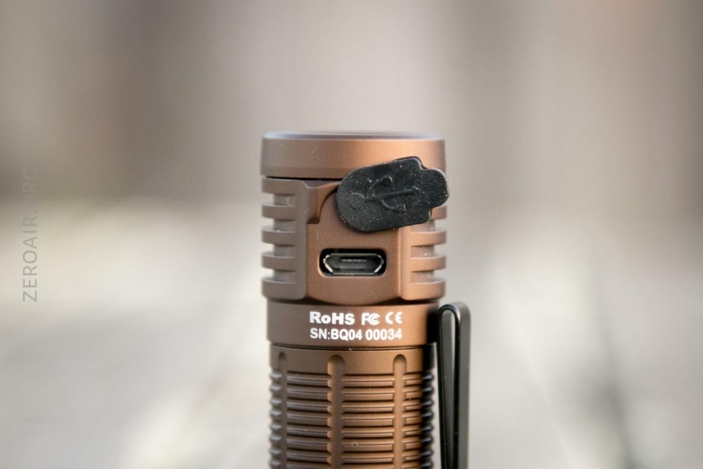Close-up of the Thrunite T1 Military Tan Flashlight standing upright, showing a covered USB-C charging port, certification markings, and part of a black pocket clip. The background is blurred.