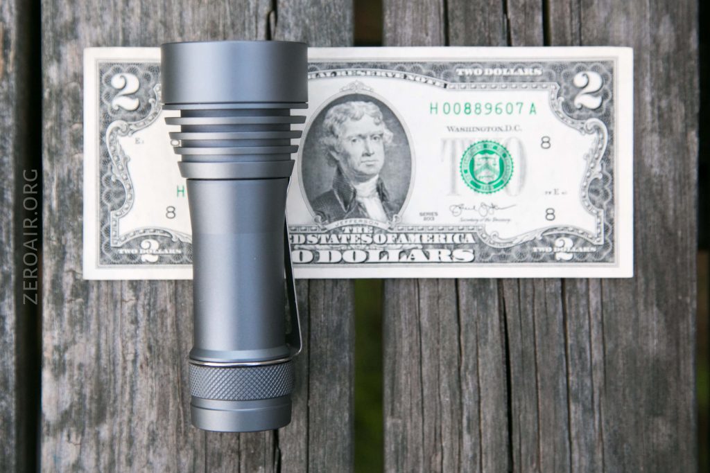 A silver flashlight is placed vertically on top of a two-dollar bill, which rests on a weathered wooden surface. The flashlight obscures the center of the bill, including part of the portrait.