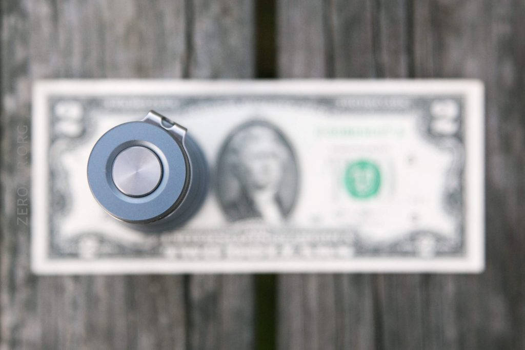 A metallic button-like object is positioned vertically above a single US one-dollar bill, which is lying flat on a wooden surface. The dollar bill is blurred and the object is in sharp focus.