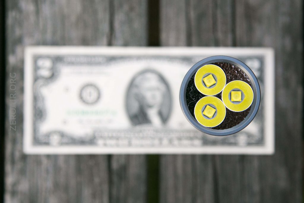 A close-up, top-down view of a flashlight with three yellow LED bulbs, placed above a blurred two-dollar bill on a wooden surface.
