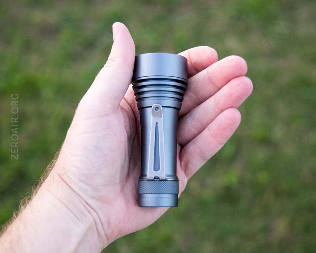 A person holds a small, metallic flashlight in their left hand against a blurred outdoor background of green grass. The flashlight is cylindrical with ridges near the top.