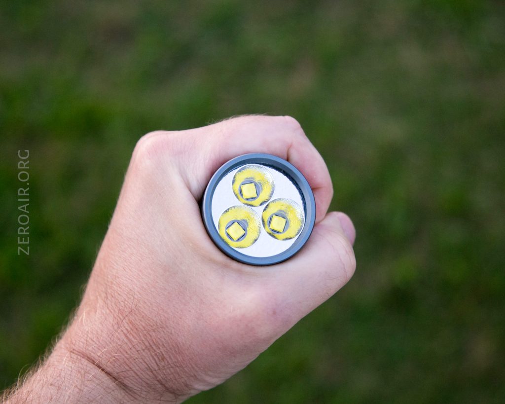 A hand holding a flashlight pointed upward, showing three yellow LED emitters arranged in a triangle on the lens, with a grassy background.
