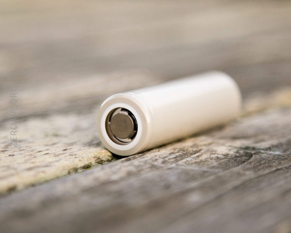A white cylindrical battery lying on its side on a weathered wooden surface, with the metal terminal facing the camera.