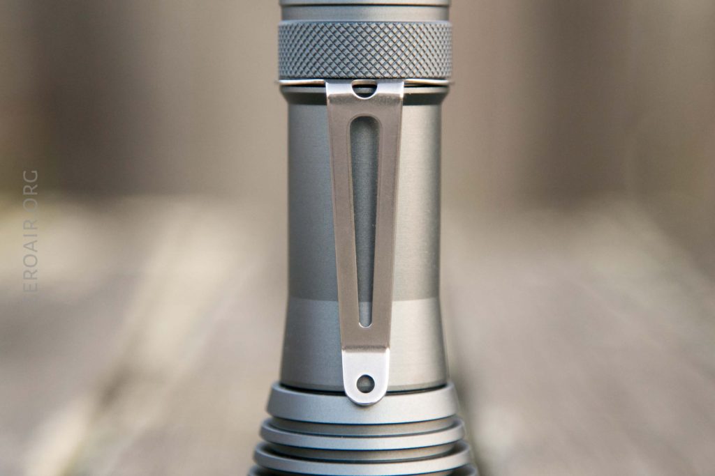 A close-up of a metallic flashlight body, showing a textured grip section and a sturdy metal pocket clip attached to the side. The background is blurred, highlighting the flashlight details.