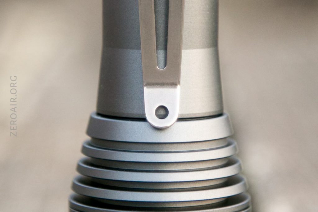 Close-up of a metallic flashlight with a sturdy pocket clip attached and horizontal cooling fins around the body. The background is out of focus, and “ZEROAIR.ORG” is faintly visible along the left side.