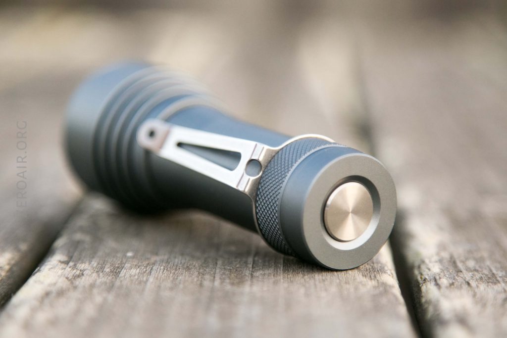 A metallic flashlight with a side clip lies on its side on a weathered wooden surface, showing the textured grip and metallic button on the end.
