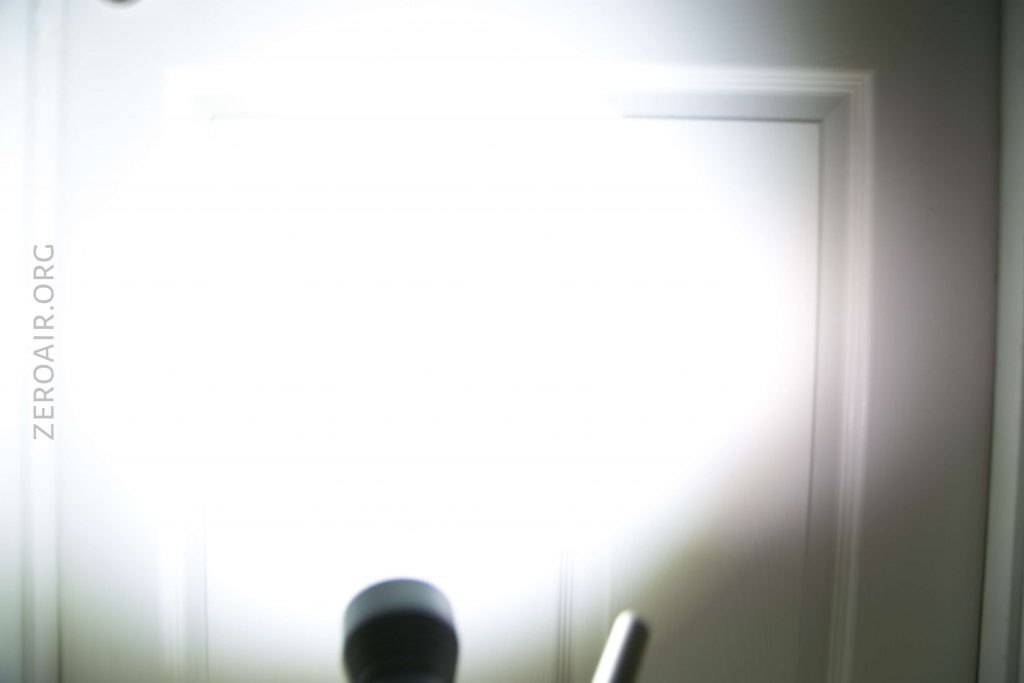 A flashlight is shining directly at a white door, creating a large, intense, circular beam of light that obscures most details of the door. The text ZEROAIR.ORG appears vertically on the left side.