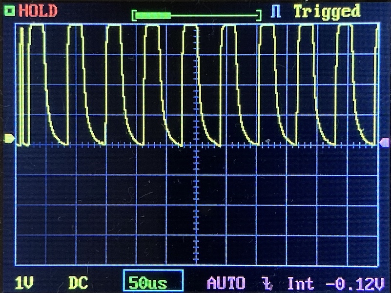 Oscilloscope screen displaying a repeating waveform with rapid peaks and exponential decay, grid lines, and measurement settings visible at the bottom, including 1V/div, 50us/div, and -0.12V offset.