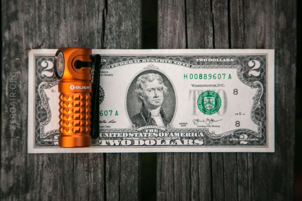 An orange Olight flashlight is placed on the left side of a two-dollar bill featuring Thomas Jefferson, both lying on a wooden surface.