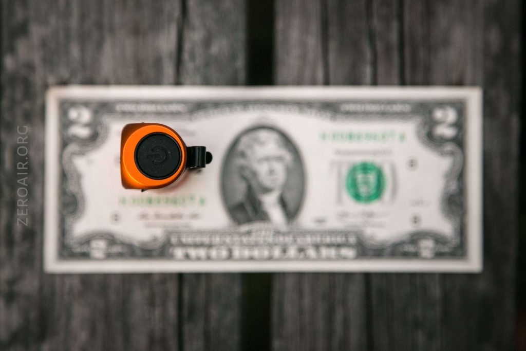 A small orange flashlight with a black power button is standing upright on the center of a U.S. two-dollar bill placed on a wooden surface. The image is seen from above.