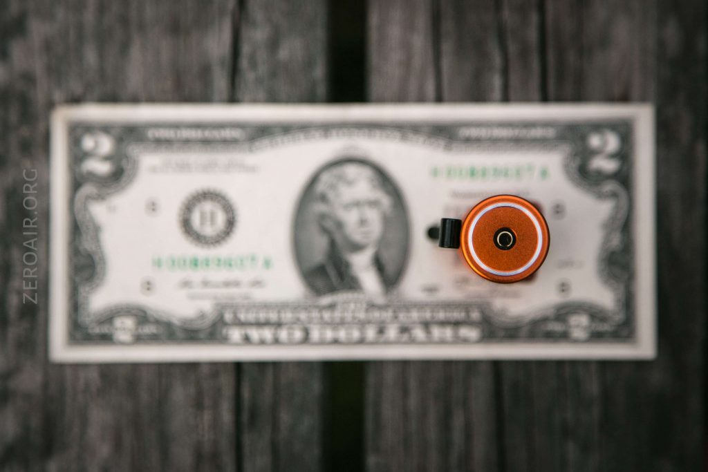 A two-dollar bill lies flat on a wooden surface with a small cylindrical object, viewed from above, placed vertically near the center of the bill. The object partially obscures the face on the bill.