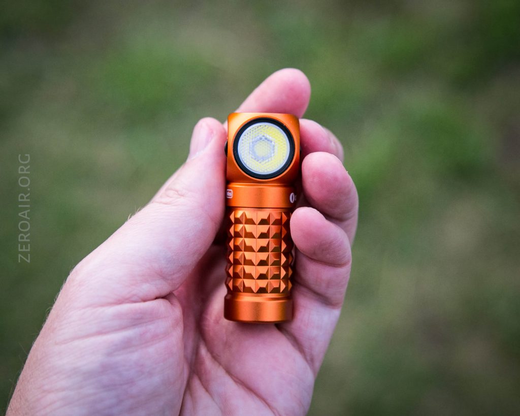 A hand holding a small, orange, textured flashlight with a side switch and a visible LED lens, against a blurred green background.