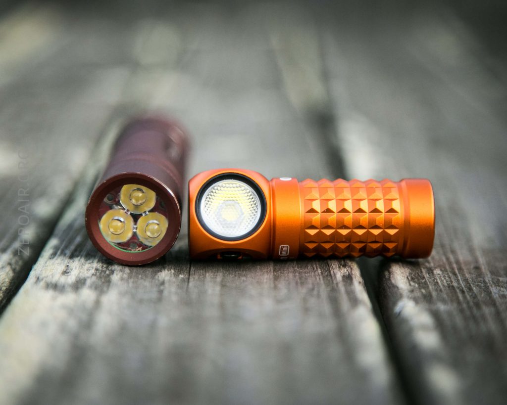 Two compact flashlights with textured orange bodies are placed on a weathered wooden surface. One flashlight is standing upright, showing three LED bulbs, while the other is lying down, displaying its lens.