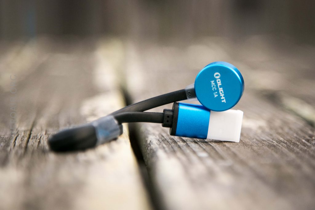 A close-up of an Olight MCC 1A magnetic charging cable with a blue and white connector placed on a weathered wooden surface. The background is blurred.