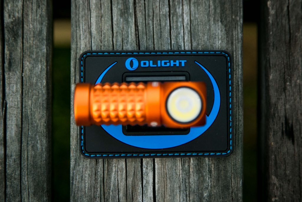An orange flashlight rests on a rectangular black and blue Olight-branded patch, placed on a weathered wooden surface with visible grain and gaps between the planks.