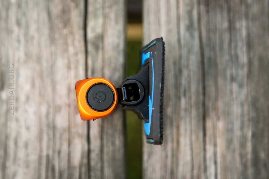 An orange and black device with a power button is clipped to a wooden surface. The background shows weathered wooden planks. The image is viewed from above.
