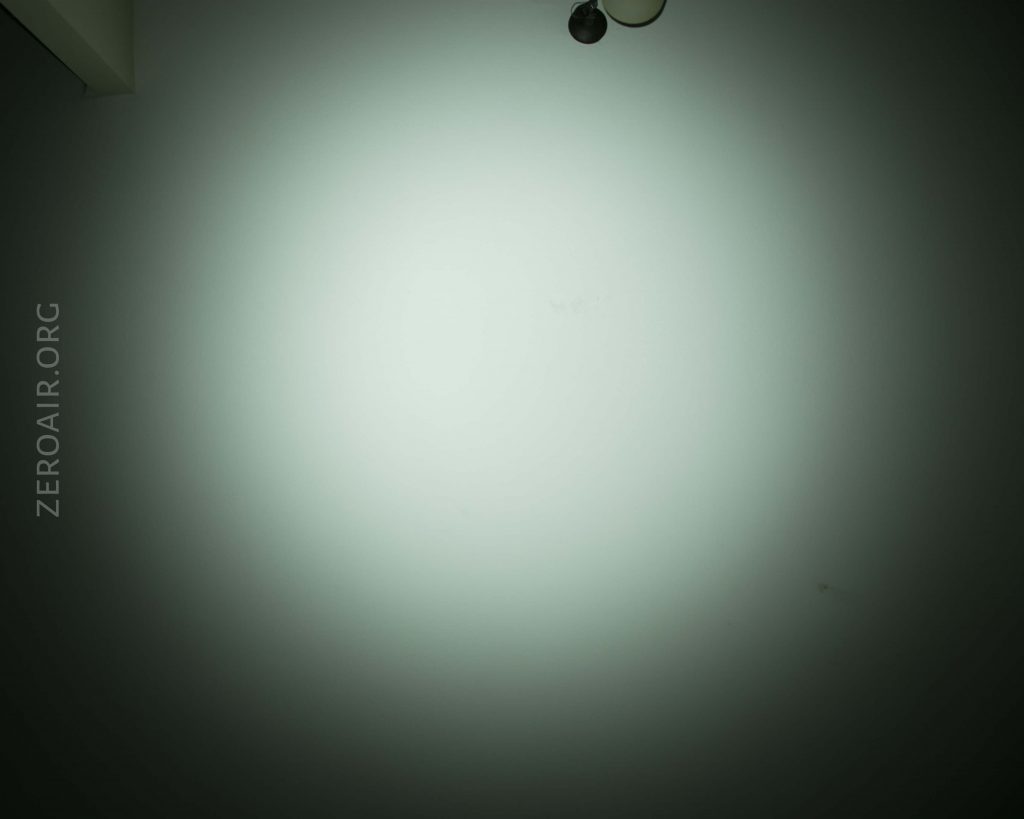 A dimly lit white wall with a small round object and part of a fixture at the top. The words ZEROAIR.ORG appear vertically on the left side of the image. The lighting creates a circular gradient on the wall.