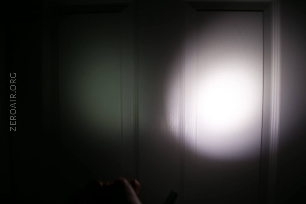 A bright flashlight beam shines on a closed white door in a dark room, creating a circular patch of light with a faintly visible hand holding the flashlight at the bottom. The text ZEROAIR.ORG appears vertically on the left side.