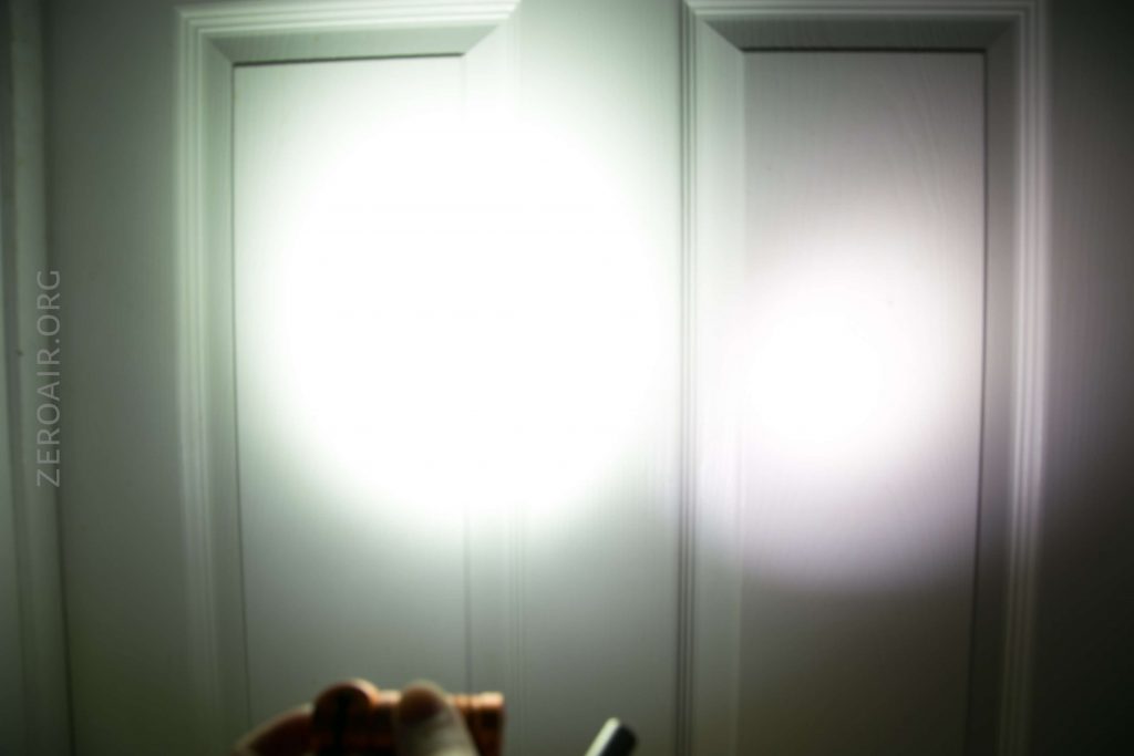 A white door with two rectangular panels is illuminated by two differently sized beams of light; a hand holding a flashlight is partially visible at the bottom of the image. The text ZEROAIR.ORG appears on the left side.