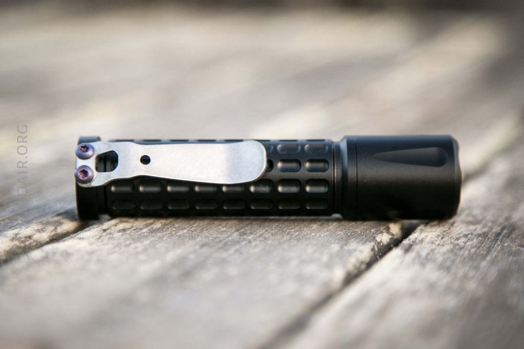 A black flashlight with a textured grip and a metal pocket clip is lying on a wooden surface. The flashlight is positioned horizontally, with the lens facing to the right.