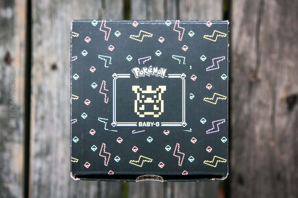 A black box features an 8-bit Pikachu graphic, the Pokémon logo, and the text BABY-G on the lid. The box is decorated with colorful patterns and is placed on a weathered wooden surface.