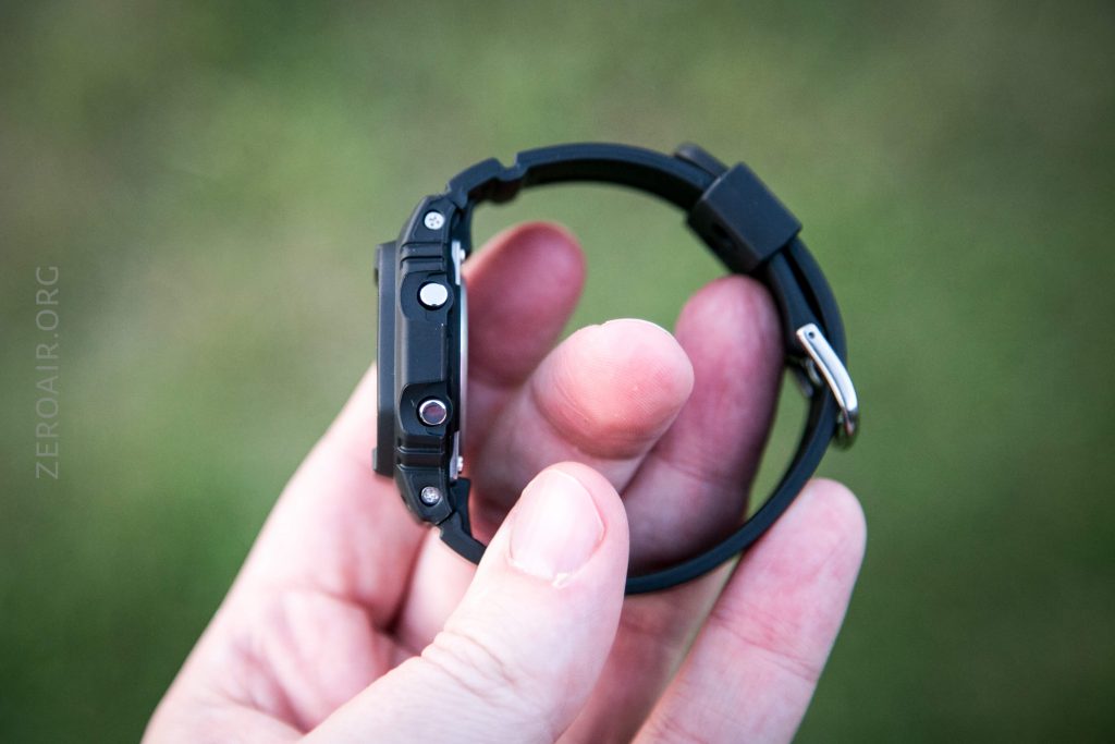 A hand holds a black wristwatch with a rubber strap, shown from the side to display its buttons and thickness. The background is blurred green grass.