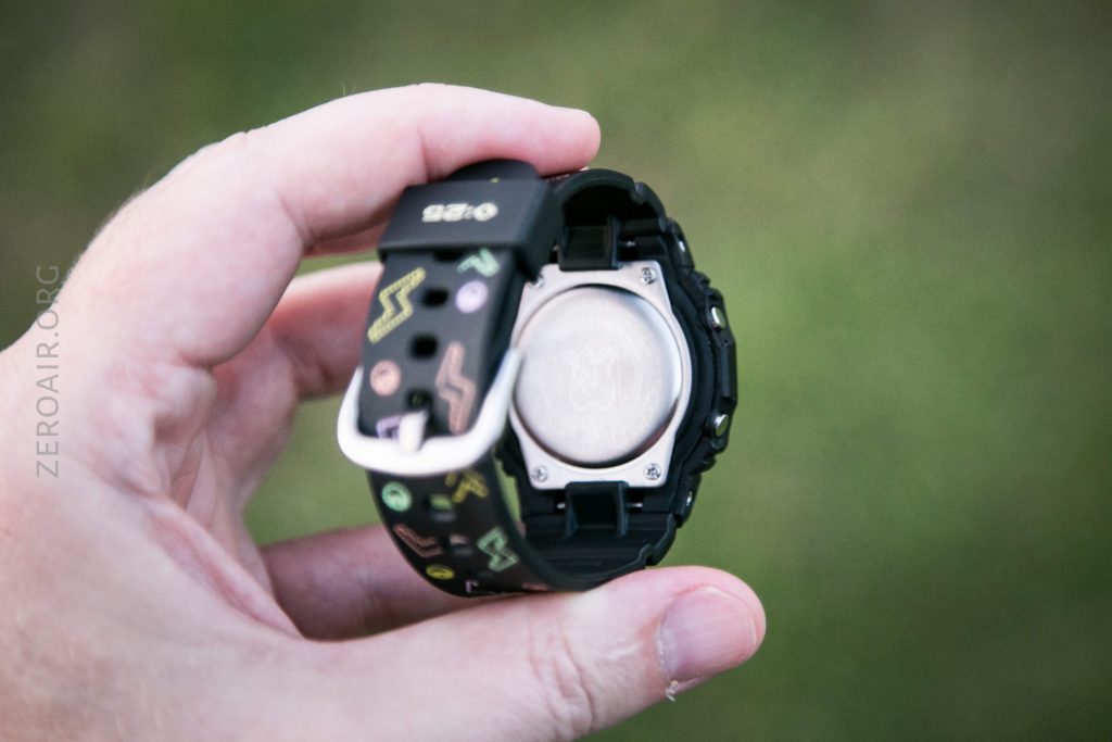 A hand holds a black digital watch with a patterned strap, showing the metal back of the watch case. The background is blurred and green. The word “ZEROAIR.ORG” is visible along the left side of the image.