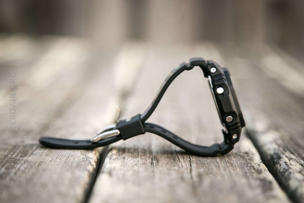 A black digital wristwatch with a thick case is positioned upright on a weathered wooden surface, showing the side profile with buttons and strap visible. The background is blurred.