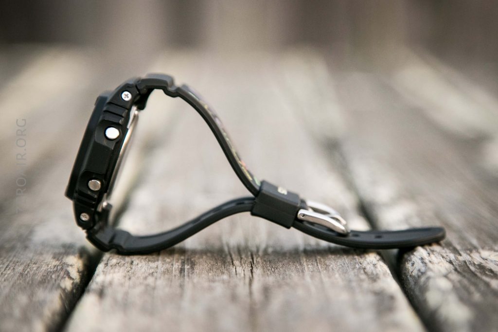 A black digital wristwatch with a curved band is placed on its side on a weathered wooden surface. The focus is on the watch, and the background is blurred.