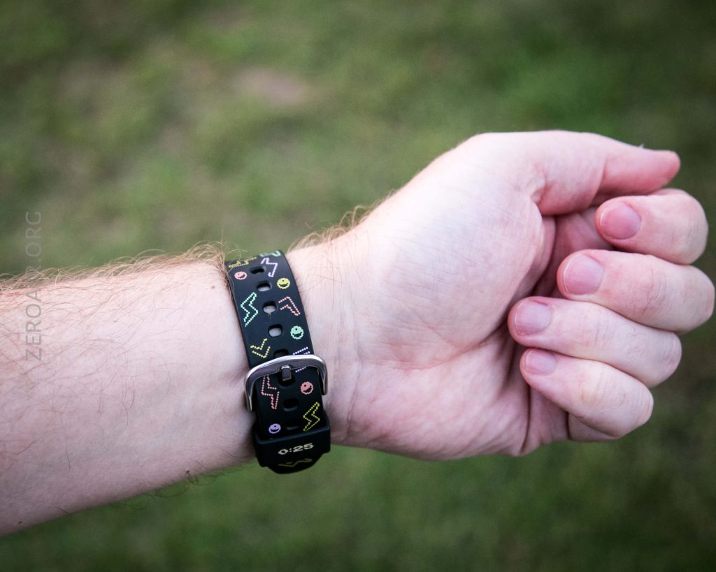 A person’s left wrist wearing a black wristband with colorful geometric shapes and symbols; the hand is relaxed and the background is grassy and blurred.
