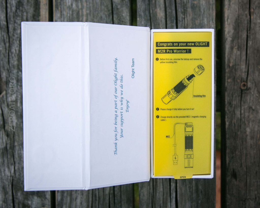 An open box on a wooden surface reveals a card with a thank you message on the left and a yellow instruction sheet with images and steps for using an Olight M2R Pro Warrior flashlight on the right.