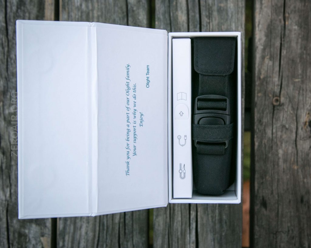 An open white box on a wooden surface contains a black fabric pouch and grooming tools. The left inner lid displays a printed message in blue text.