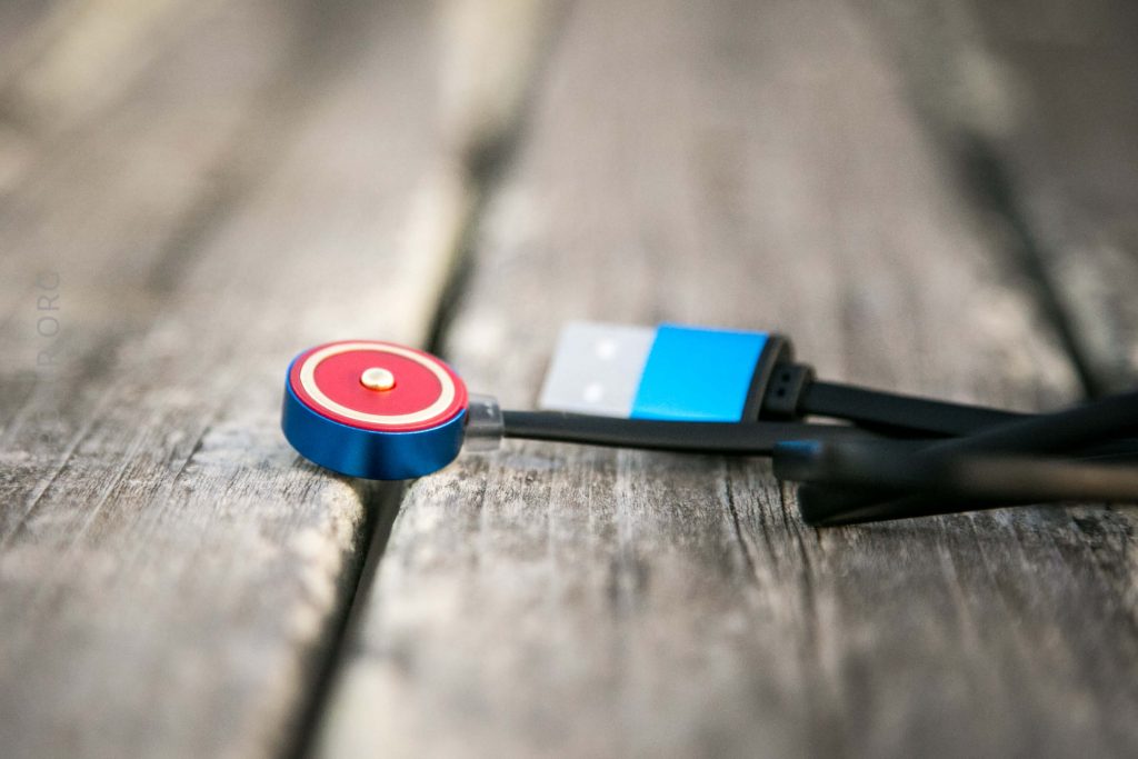 A close-up of a blue and red magnetic USB charging cable with a USB-A connector, lying on a weathered wooden surface. The cable is partially coiled and the connector is in focus.