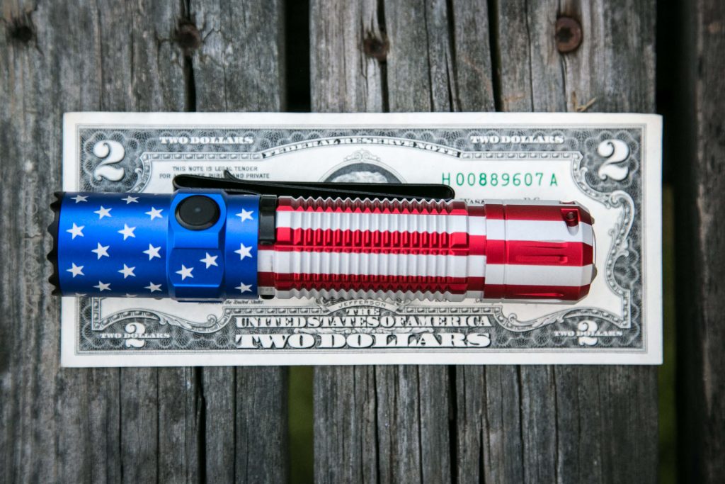 A flashlight with an American flag design lies on top of a two-dollar bill on a weathered wooden surface.