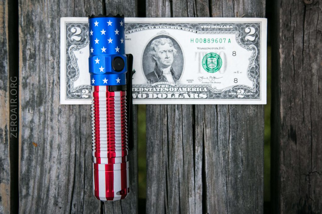 A red, white, and blue flashlight with a stars and stripes pattern lies vertically on a rustic wooden surface, placed in front of a two-dollar bill featuring Thomas Jefferson.