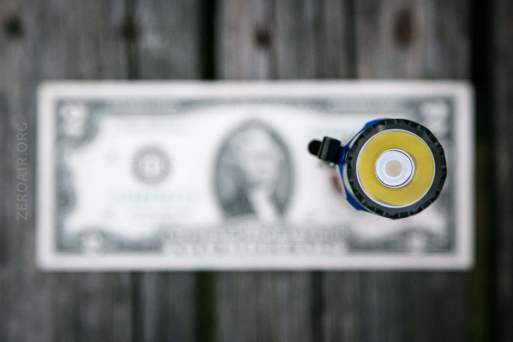 A single dollar bill lies flat on a wooden surface, with the top view of a cylindrical flashlight and its battery centered above the bill, partly obscuring it.