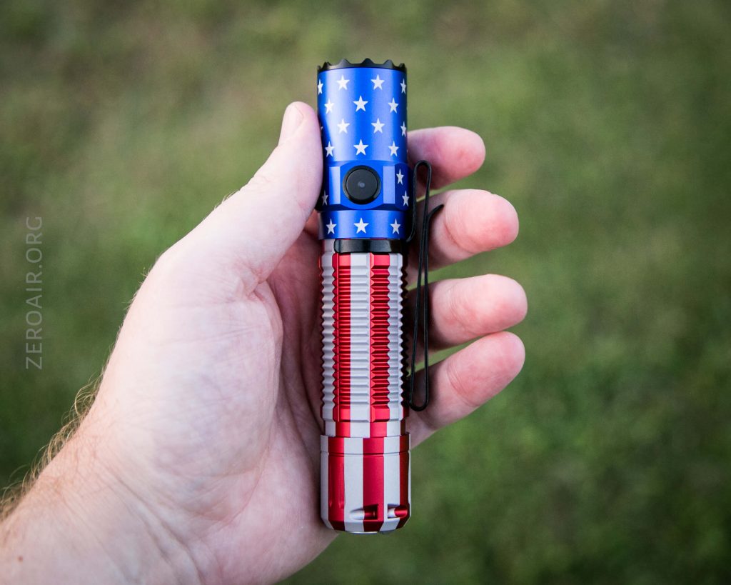 A hand holds a flashlight designed with an American flag pattern, featuring blue with white stars on the top and red and white stripes on the bottom, against a blurred grassy background.