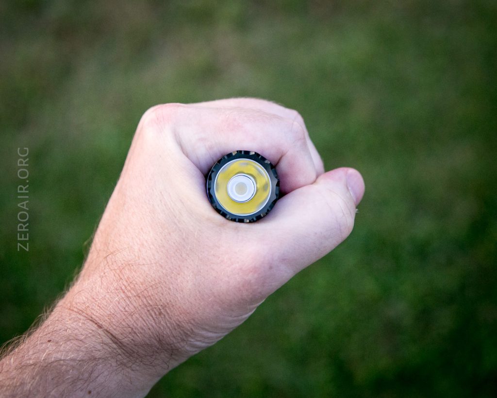 A hand holds a cylindrical object horizontally, viewed from above so that the round, yellow end with a white center is visible. The background is a blurred grassy surface.