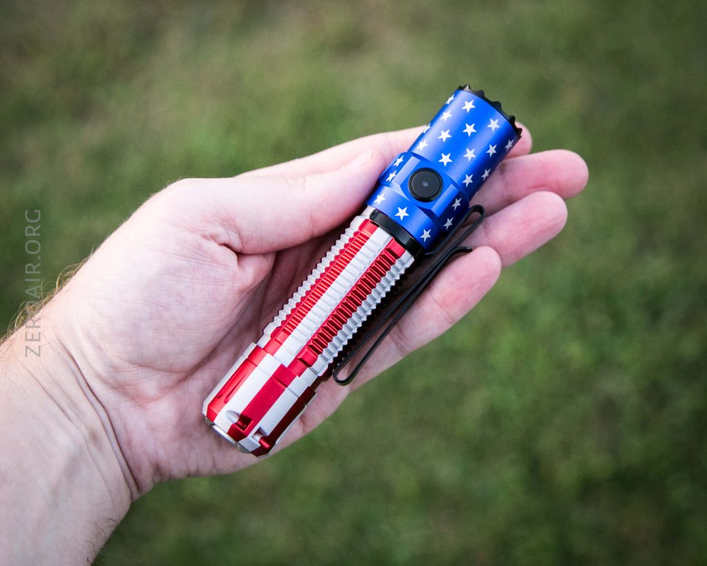 A person holds a flashlight decorated with an American flag design, featuring blue with white stars and red and white stripes, against a blurred green background.