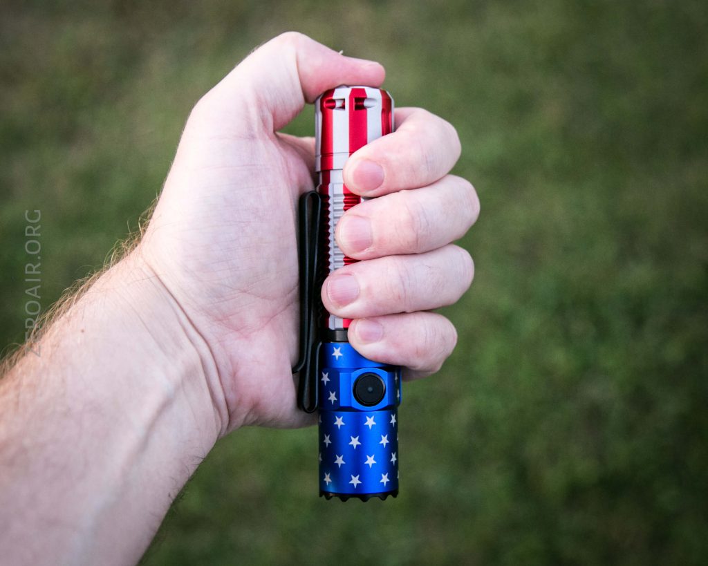 A hand holding a flashlight with a red top, silver middle, and blue bottom decorated with white stars. The background is green and blurry grass.